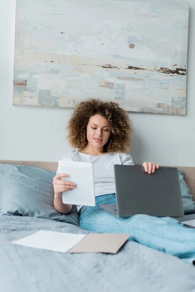 Donna sorridente con computer portatile e tablet digitale seduta sul letto vicino ai documenti — Foto stock