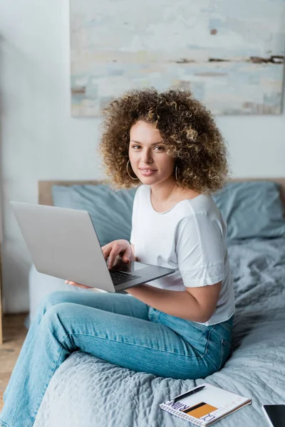 Mulher encaracolado sorrindo para a câmera enquanto sentado na cama com laptop — Fotografia de Stock