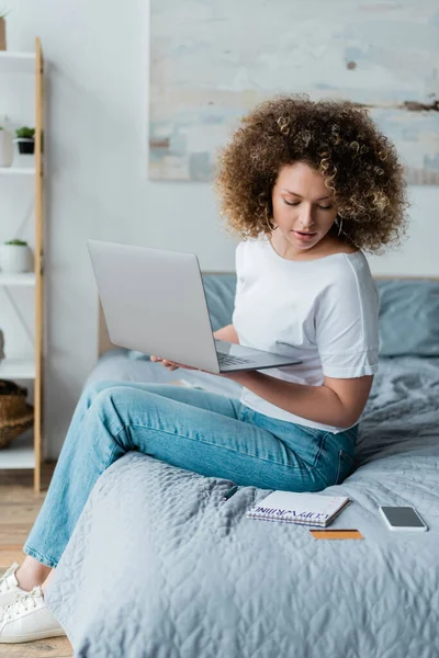 Lockige Frau sitzt mit Laptop im Bett und schaut auf Notizbuch mit Schriftzug — Stockfoto