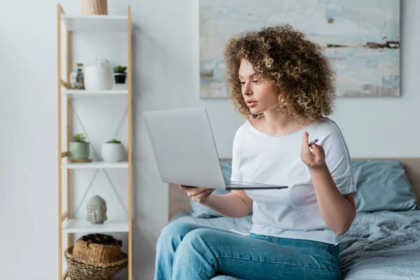 Curly woman in t-shirt and jeans sitting with laptop on bed at home — Stock Photo