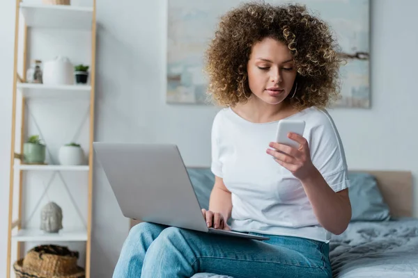 Donna riccia in t-shirt bianca seduta sul letto con smartphone e laptop — Foto stock