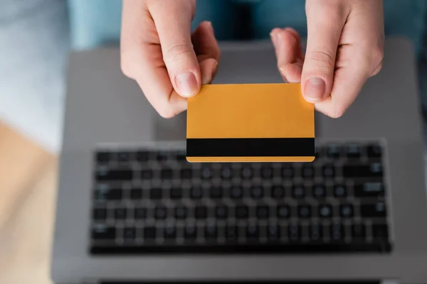 Vista superior de la tarjeta de crédito en manos de la mujer recortada cerca de la computadora portátil borrosa - foto de stock