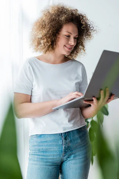 Mujer sonriente con el pelo ondulado de pie con el ordenador portátil en primer plano borrosa - foto de stock