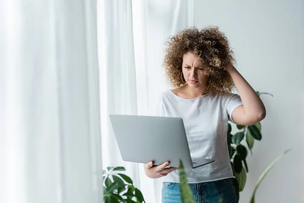 Nachdenkliche Frau berührt welliges Haar, während sie zu Hause auf Laptop schaut — Stockfoto