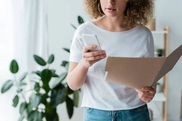 Partial view of woman in white t-shirt holding smartphone and folder at home — Stock Photo