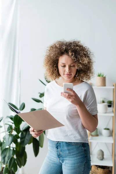 Femme bouclée tenant dossier avec des documents tout en utilisant un téléphone portable à la maison — Photo de stock