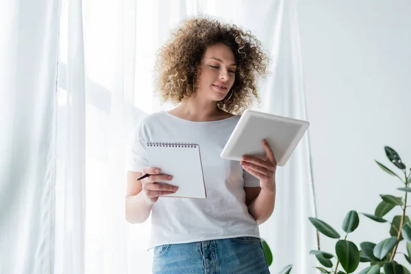 Femme bouclée souriant tout en se tenant debout avec tablette numérique et ordinateur portable près du rideau blanc — Photo de stock