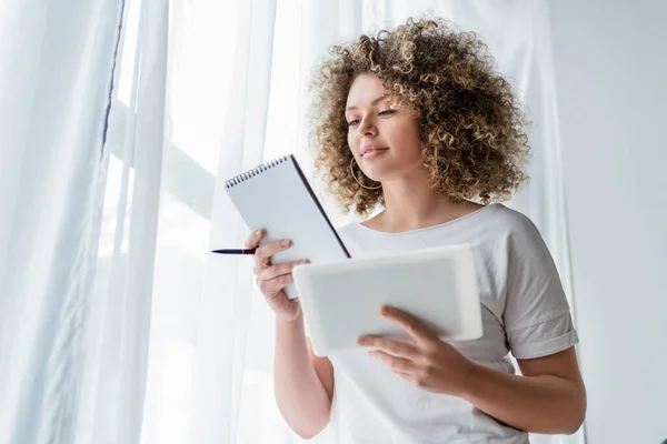 Junge lächelnde Frau steht mit Notizbuch und digitalem Tablet neben weißem Vorhang — Stockfoto