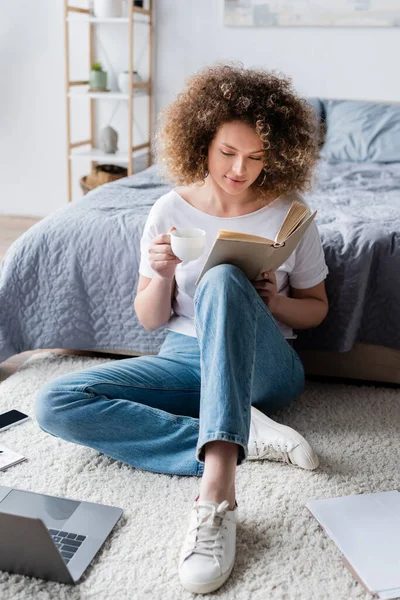 Jeune femme avec tasse de café livre de lecture sur le sol près de l'ordinateur portable — Photo de stock