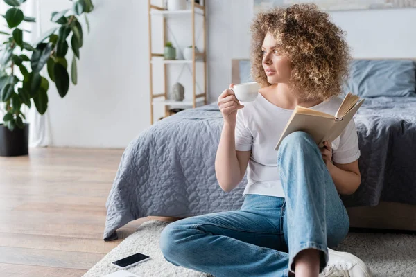 Lockige Frau mit Buch und Kaffeetasse sitzt auf dem Boden im Schlafzimmer — Stockfoto