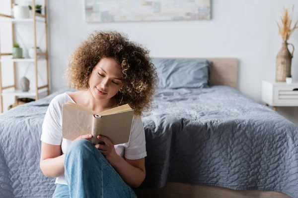 Pleased woman with curly hair sitting near bed and reading book — Stock Photo