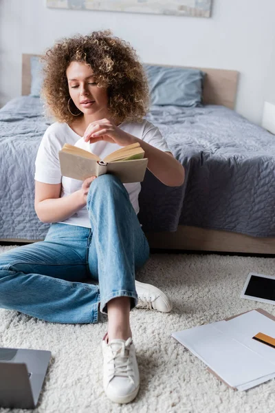 Longitud completa de la mujer en jeans libro de lectura en el suelo cerca de los papeles y el ordenador portátil - foto de stock