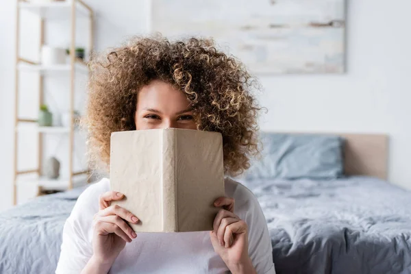 Curly woman with cheerful eye expression obscuring face with book and looking at camera — Stock Photo