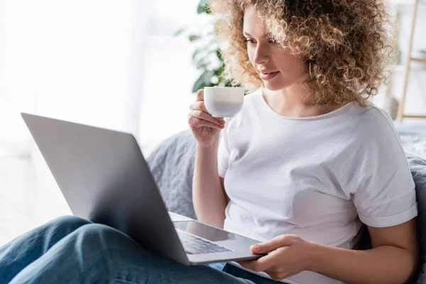 Glückliche und lockige Frau mit Kaffeetasse blickt auf Laptop im Schlafzimmer — Stockfoto