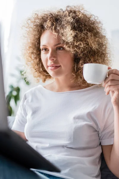 Zufriedene Frau im weißen T-Shirt mit Kaffeetasse und Blick auf verschwommenen Laptop — Stockfoto