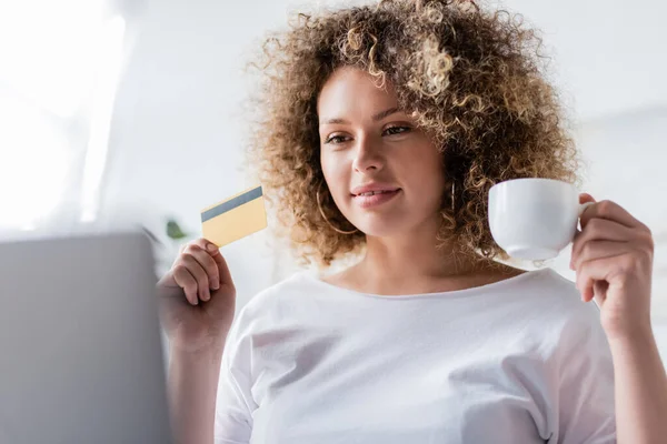 Mujer sonriente con taza de café y tarjeta de crédito mirando borrosa portátil - foto de stock