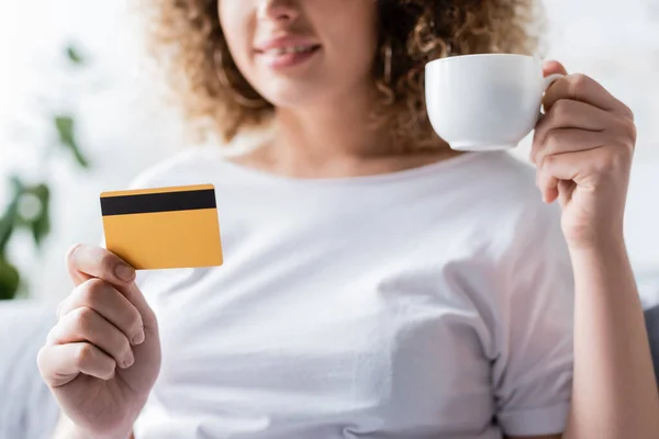 Частичный вид улыбающейся женщины, держащей чашку кофе и кредитную карту на размытом фоне — стоковое фото