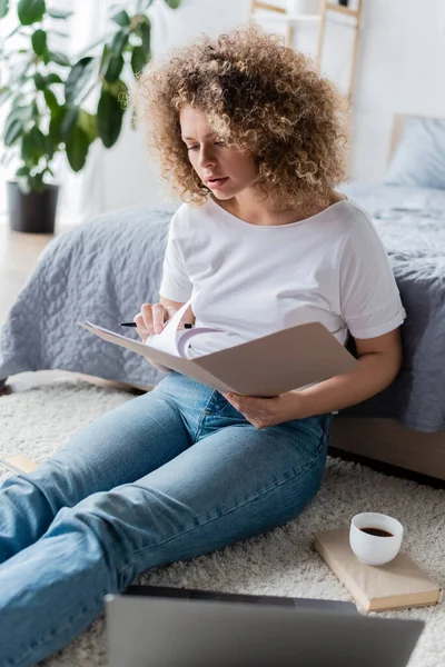 Mujer joven con documentos que trabajan en el suelo cerca de la computadora portátil y taza de café - foto de stock