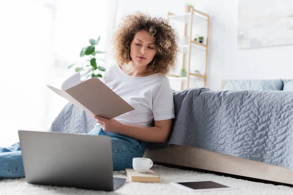 Mujer rizada con carpeta sentada en el suelo cerca de la computadora portátil y taza de café - foto de stock