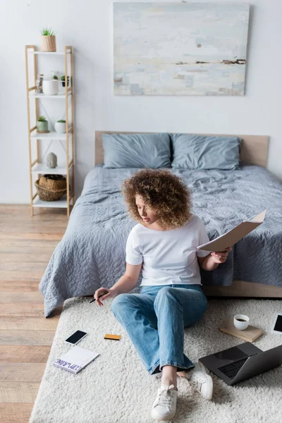 Mulher encaracolado em jeans trabalhando no chão perto de gadgets e notebook com letras copywriting — Fotografia de Stock