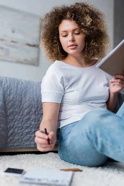 Кучерява жінка в білій футболці сидить на підлозі з ручкою і текою — стокове фото
