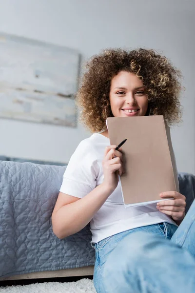 Mujer feliz con pluma y carpeta sentado en el suelo en el dormitorio y mirando a la cámara - foto de stock