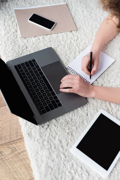 Top view of cropped woman writing in notebook near devices with blank screen on floor — Stock Photo
