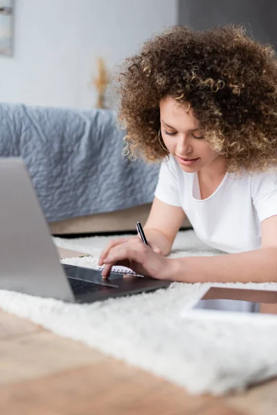 Mulher encaracolado positivo escrevendo perto laptop no chão no quarto — Fotografia de Stock