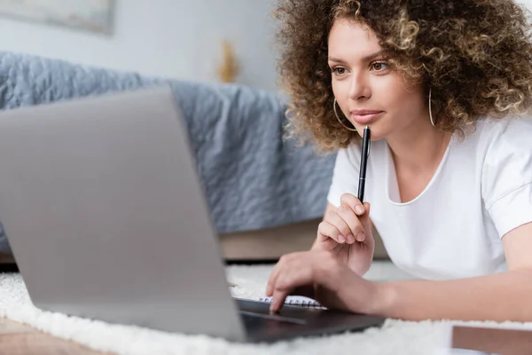 Mulher sorrindo segurando caneta e usando laptop no chão no quarto — Fotografia de Stock