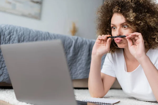 Curly woman grimacing with pen while lying near laptop on floor — Stock Photo