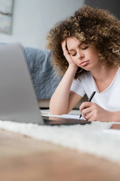 Nachdenkliche Frau schreibt in Notizbuch neben verschwommenem Laptop auf Boden im Schlafzimmer — Stockfoto