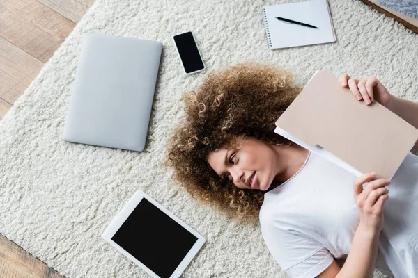 Вид сверху кудрявой женщины с папкой, лежащей на ковре рядом с устройствами и ноутбуком — стоковое фото