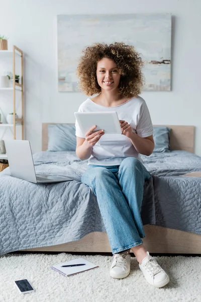 Mujer feliz con la tableta digital sonriendo a la cámara cerca del ordenador portátil en la cama - foto de stock