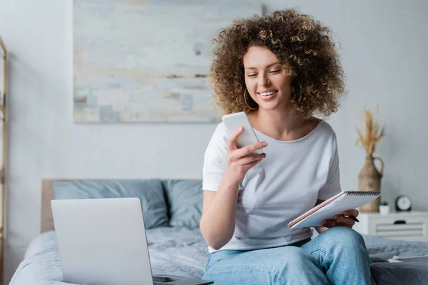 Mujer alegre usando el teléfono móvil mientras está sentado con portátil cerca de la computadora portátil - foto de stock