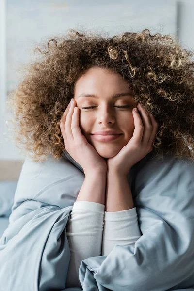 Positive Frau mit welligem Haar und Händen in Gesichtsnähe träumt mit geschlossenen Augen im Schlafzimmer — Stockfoto