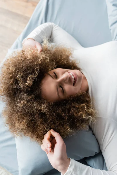 Vue de dessus de femme bouclée joyeuse avec les yeux fermés étirant sur le lit à la maison — Photo de stock