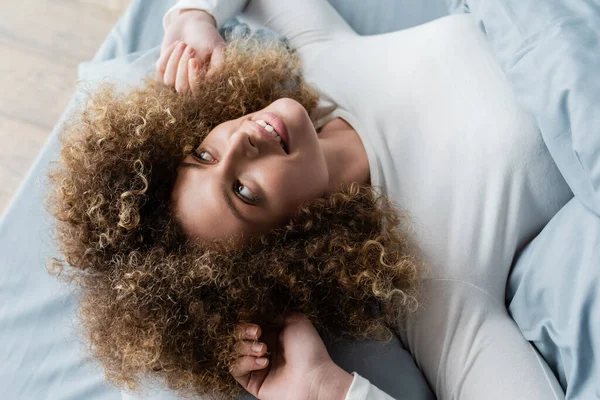 Draufsicht einer lächelnden Frau mit welligem Haar, die auf dem Bett liegt und wegschaut — Stockfoto