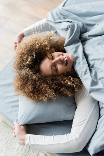 Vista superior da mulher encaracolada sorridente com os olhos fechados esticados na cama em casa — Fotografia de Stock