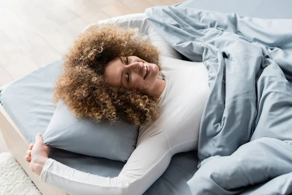 Vista de ángulo alto de la mujer alegre con el pelo ondulado acostado en la ropa de cama gris con los ojos cerrados - foto de stock