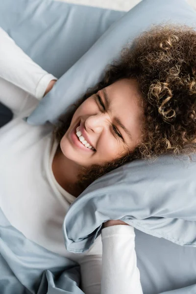 Draufsicht der fröhlichen Frau, die Ohren mit Kissen auf dem Bett bedeckt — Stockfoto