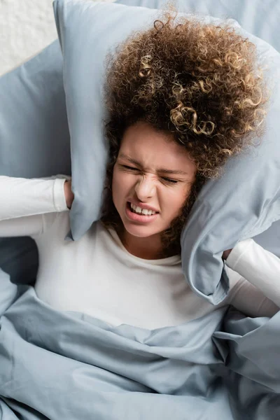 Top view of displeased woman covering ears with pillow while lying on bed — Stock Photo