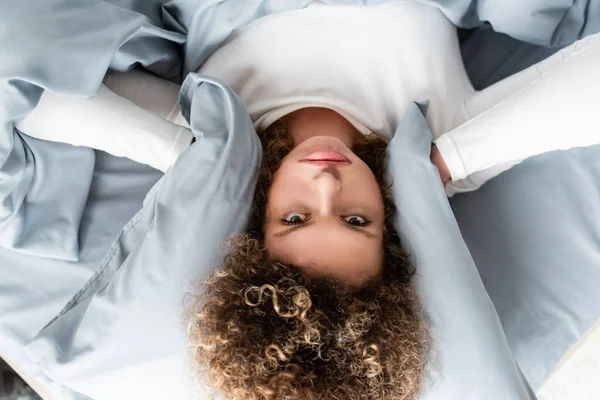 Vista superior de mujer disgustada cubriendo las orejas con almohada y mirando a la cámara - foto de stock