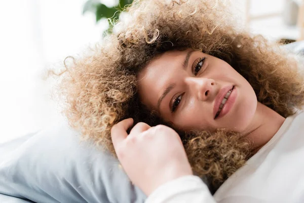 Довольная молодая женщина с волнистыми волосами лежит на подушке и улыбается в камеру — стоковое фото