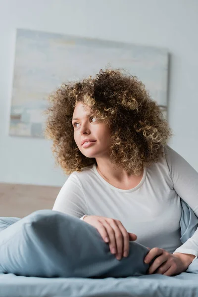 Junge Frau mit welligem Haar lächelt und schaut im Schlafzimmer weg — Stockfoto