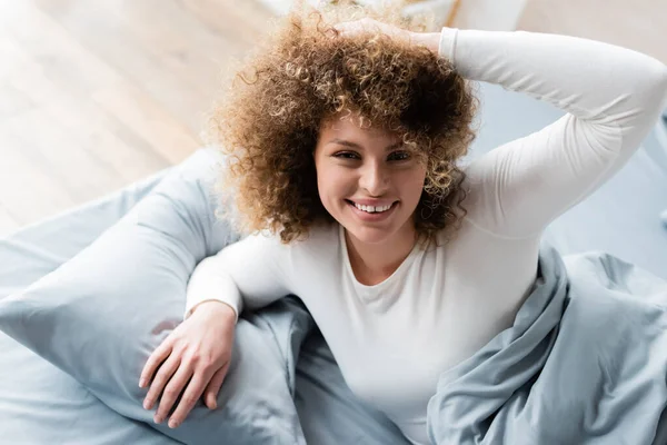 Молодая женщина трогает кудрявые волосы и улыбается в камеру на кровати дома — стоковое фото