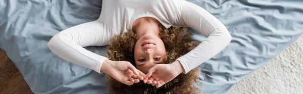 Top view of cheerful woman with curly hair lying on grey bedding, banner — Stock Photo