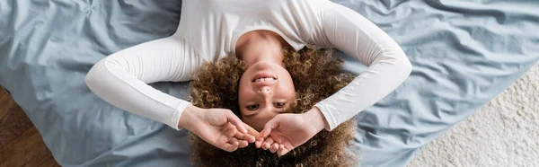 Draufsicht auf fröhliche Frau mit lockigem Haar, die auf grauem Bettzeug liegt, Banner — Stockfoto