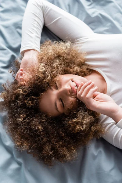 Vue de dessus de la femme bouclée souriante tenant la main près du visage tout en étant couché sur le lit — Photo de stock