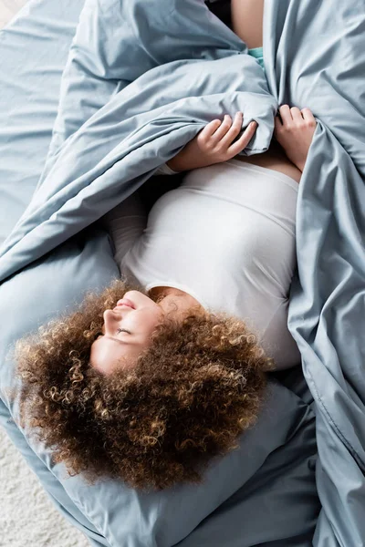 Visão de alto ângulo de mulher encaracolada com olhos fechados deitado sobre cama cinza — Fotografia de Stock