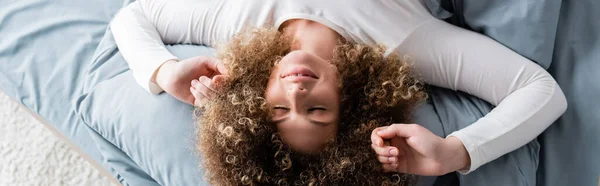 Вид зверху задоволеної жінки з кучерявим волоссям, що розтягується на ліжку вдома, банер — стокове фото